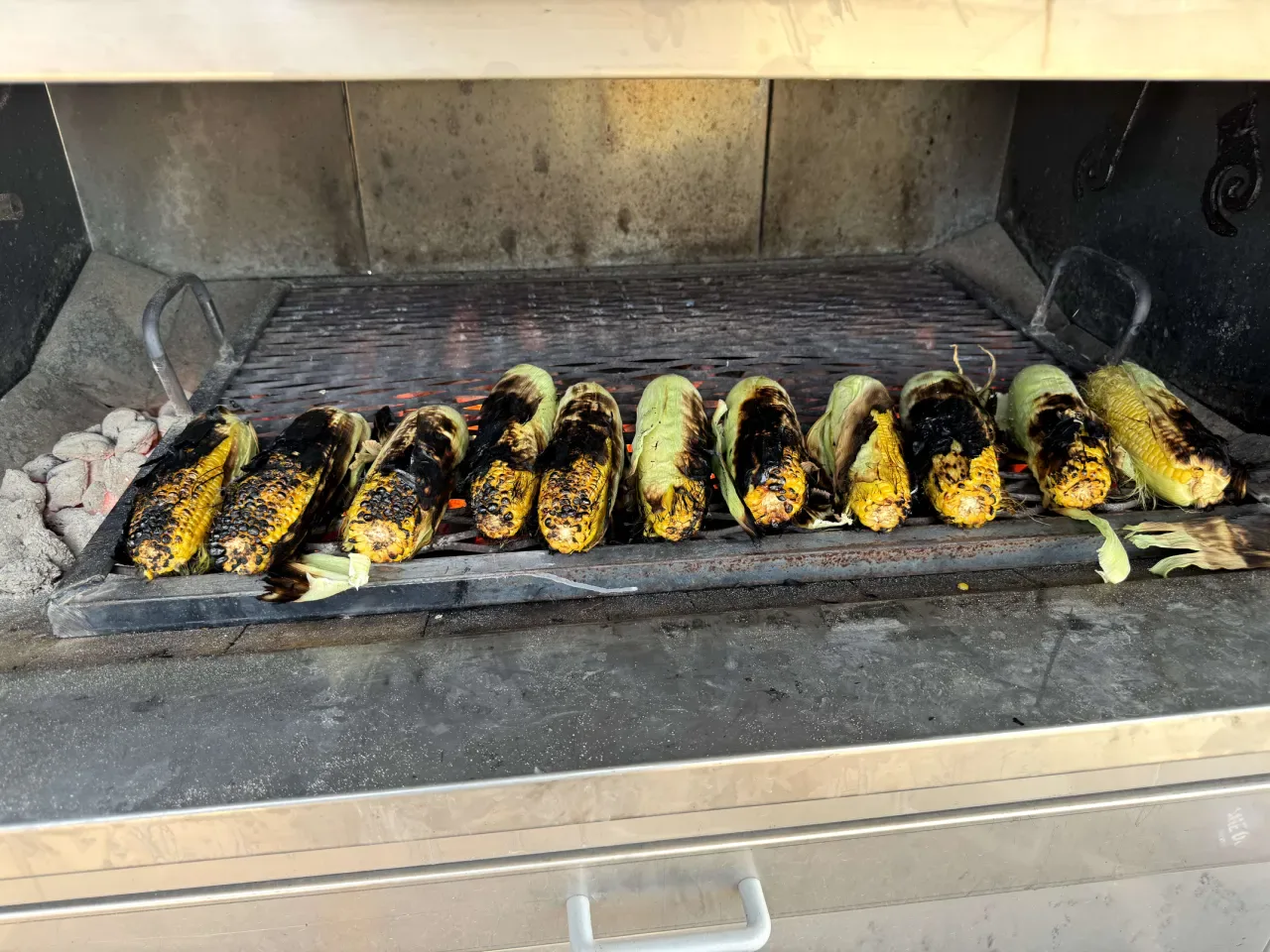 A row of elote charring on the grill over hot coals